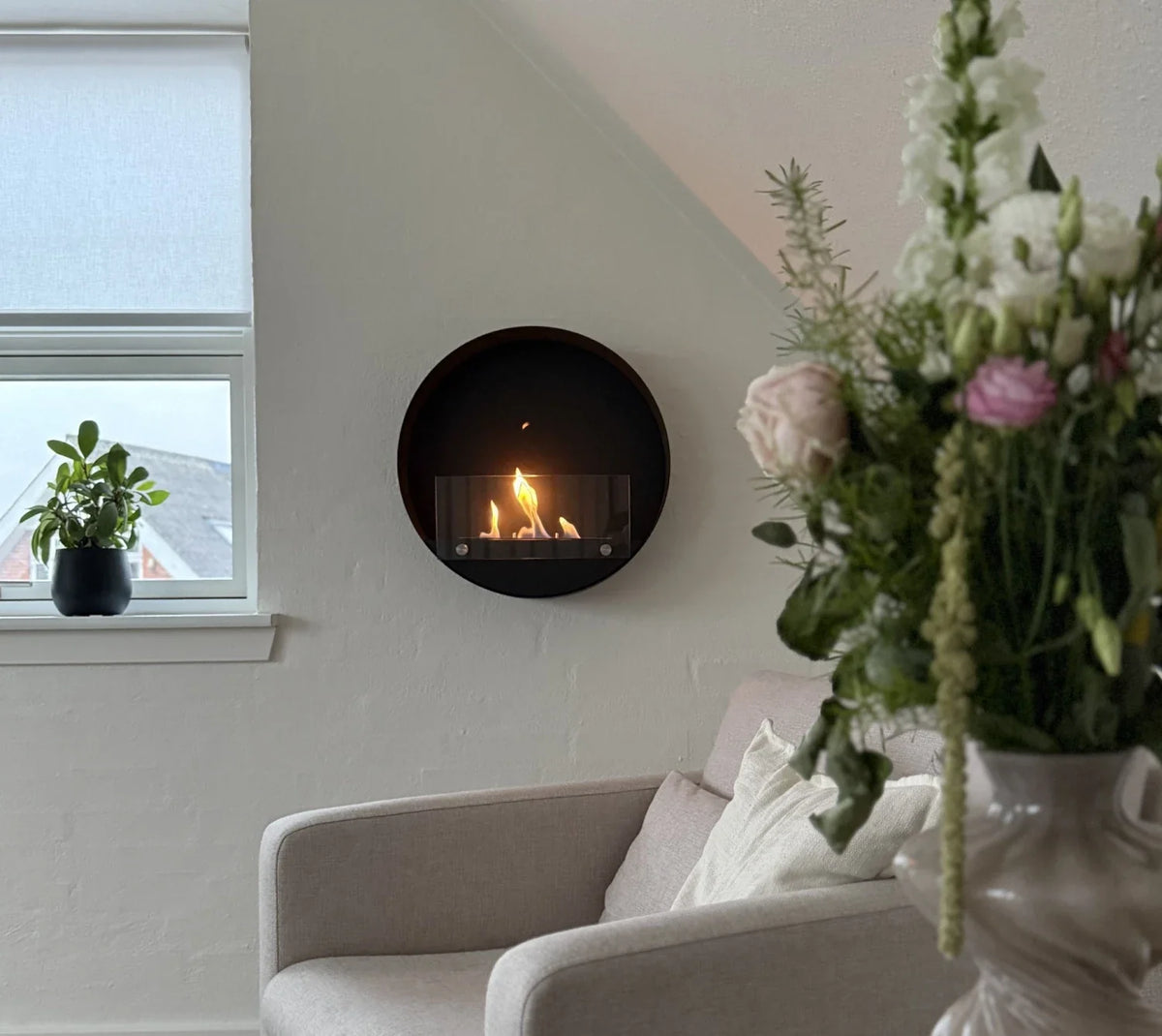 Delaware S black round bio fireplace mounted in a bright attic-style living room with plants and flowers, lit with a realistic flame
