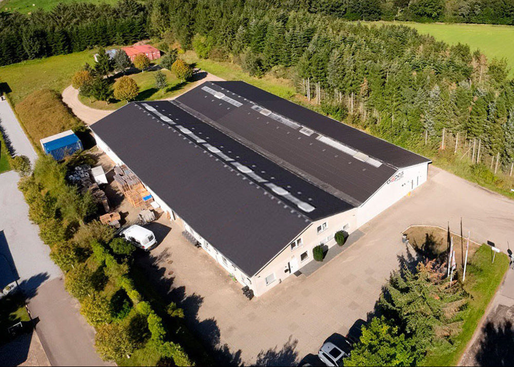 Aerial view of a large rectangular building with a dark gray roof and white walls, surrounded by green fields, trees, and a paved yard with parked cars.